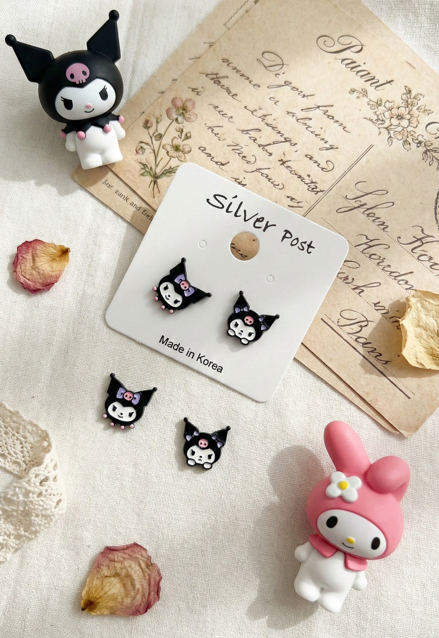 Flat lay of Kuromi character earrings on vintage postcard with dried flowers and Sanrio toys