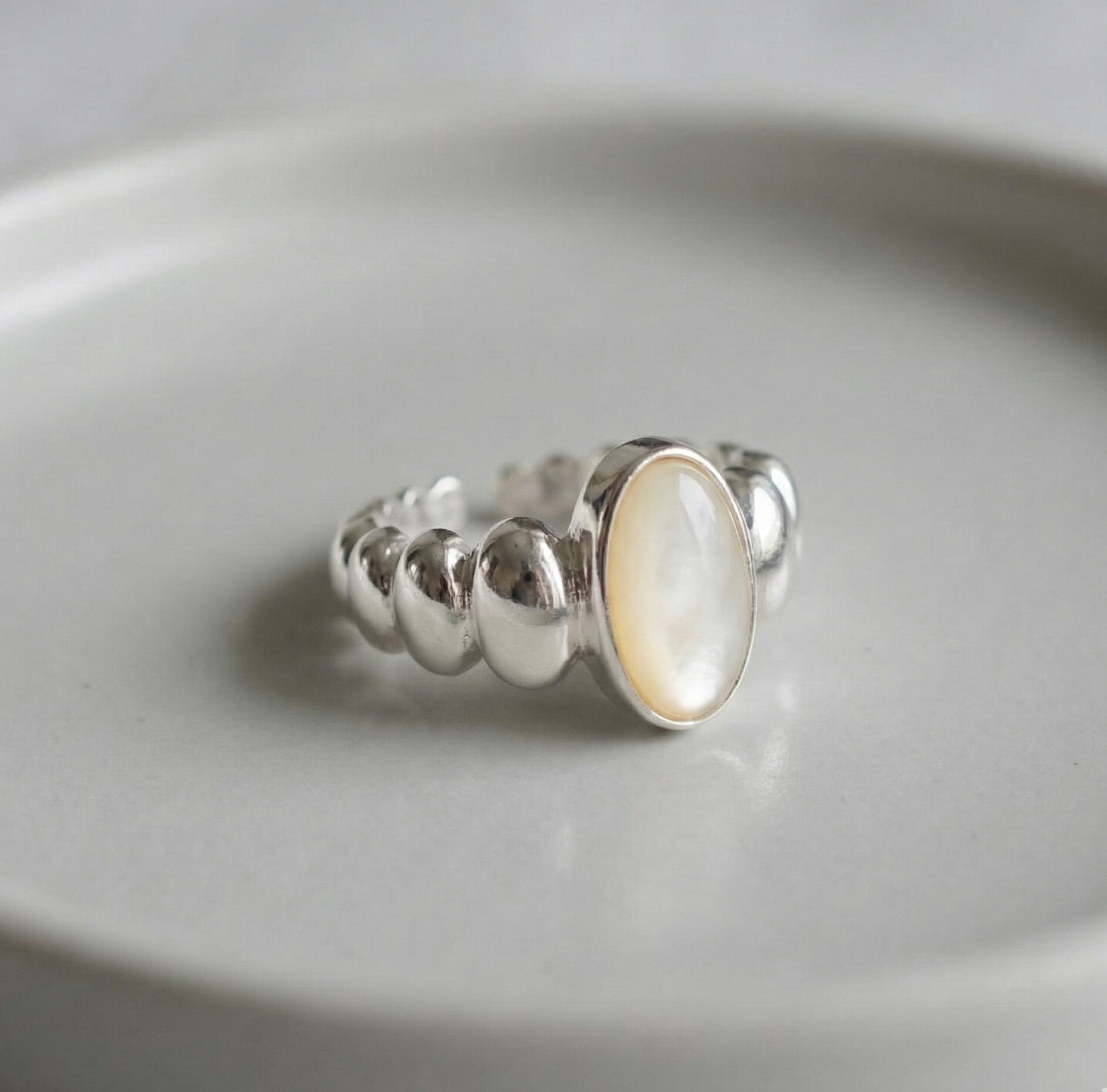 Close-up detail of 925 sterling silver mother of pearl oval ring with bold beaded ball band on white ceramic plate showing iridescent pearl stone and high-polish silver finish