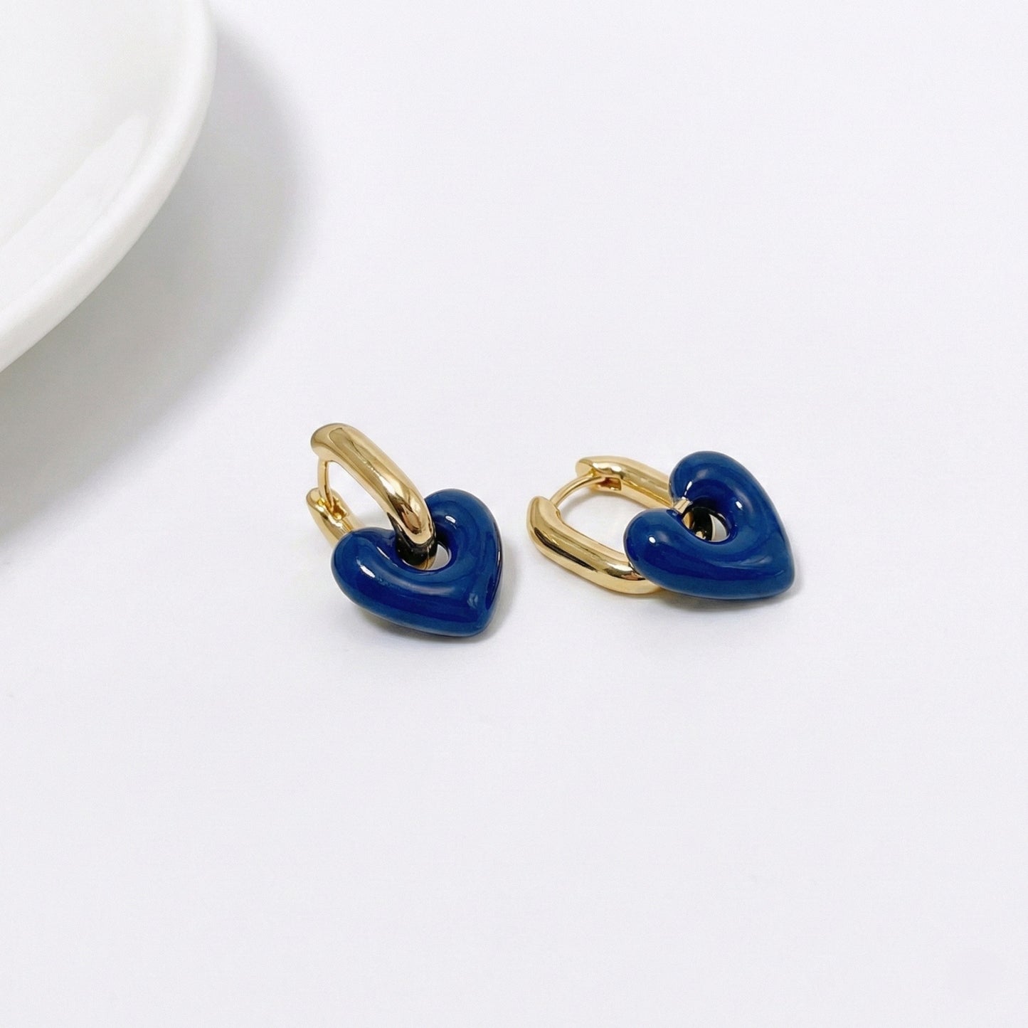 Navy blue puffy heart earrings hanging from gold huggie hoops shown on white background