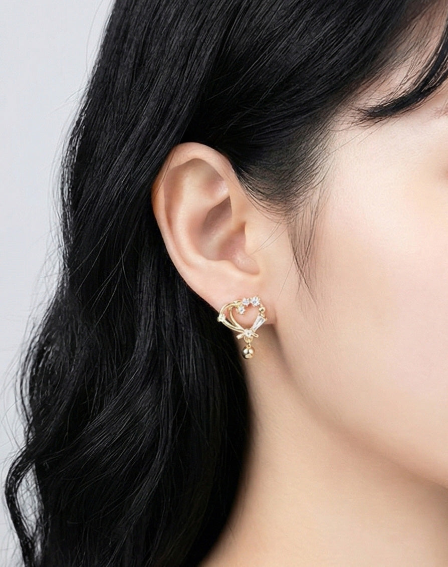 Side view of woman wearing gold heart earrings with CZ stones and drop ball charm, showing elegant romantic design, brass with 925 silver posts, made in Korea
