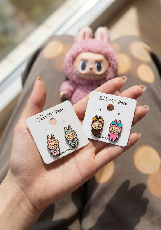 Hand holding two white earring cards with Labubu pink and brown bunny earrings with purple Labubu plush toy in background