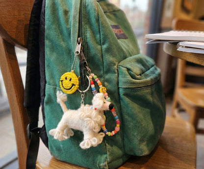 Fluffy white felt poodle bag charm with beaded keychains and smiley face charm on a green canvas backpack