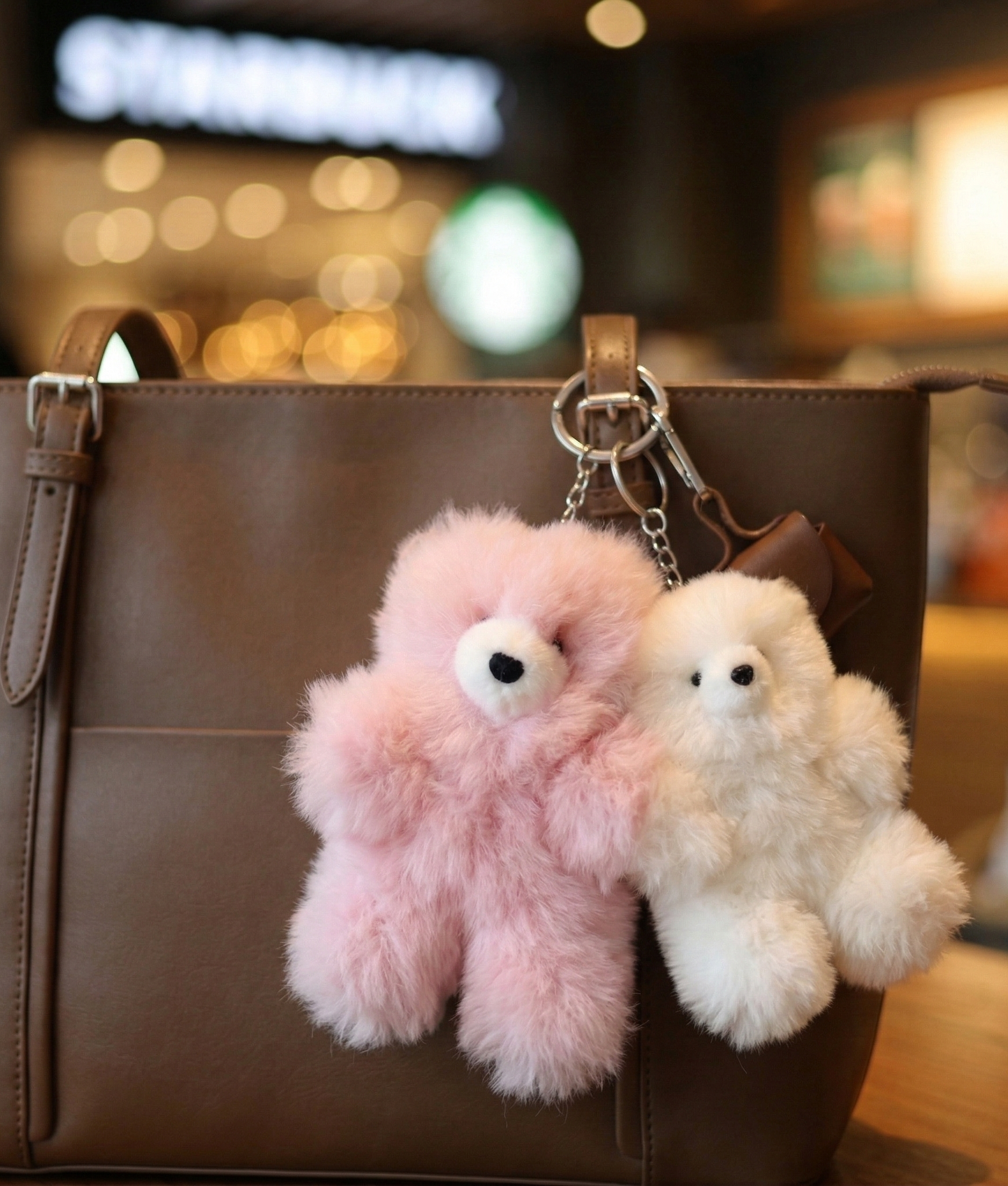 Pink and white fluffy alpaca bear bag charms hanging from a brown leather tote bag handle in a bokeh cafe background
