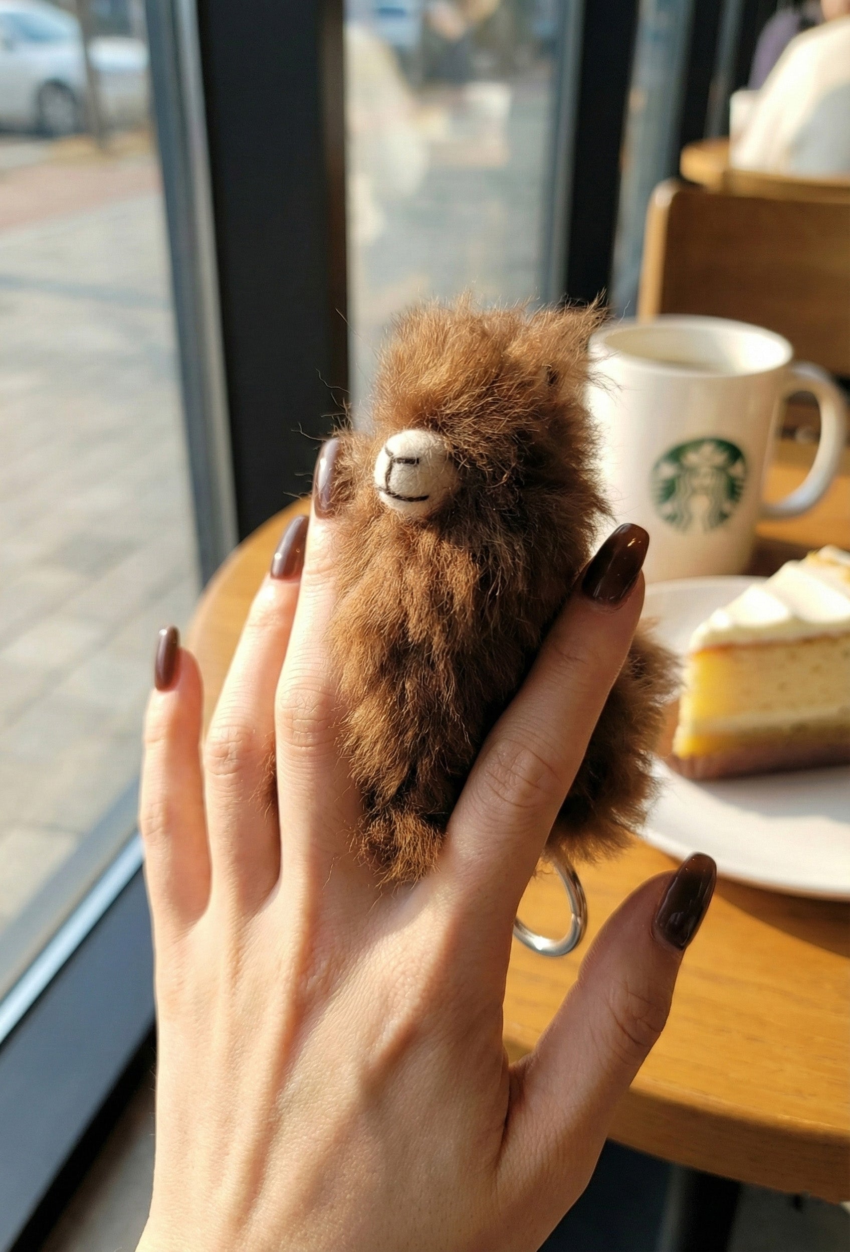 Hand holding soft alpaca plush keychain at cafe table with coffee cup in background