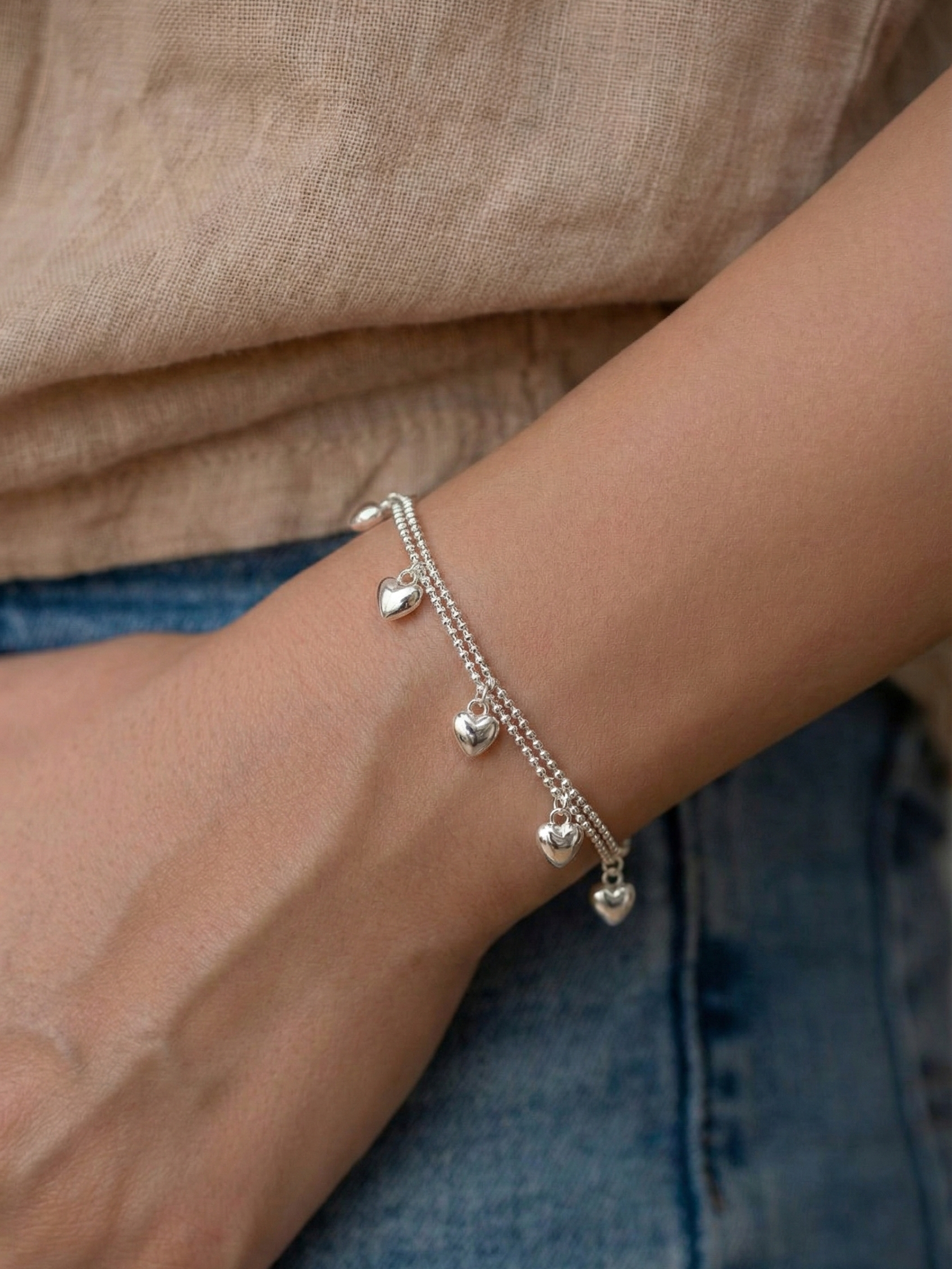 Close-up of a dainty 925 silver heart charm bracelet worn on a wrist against beige linen top and blue denim jeans, casual everyday layered style