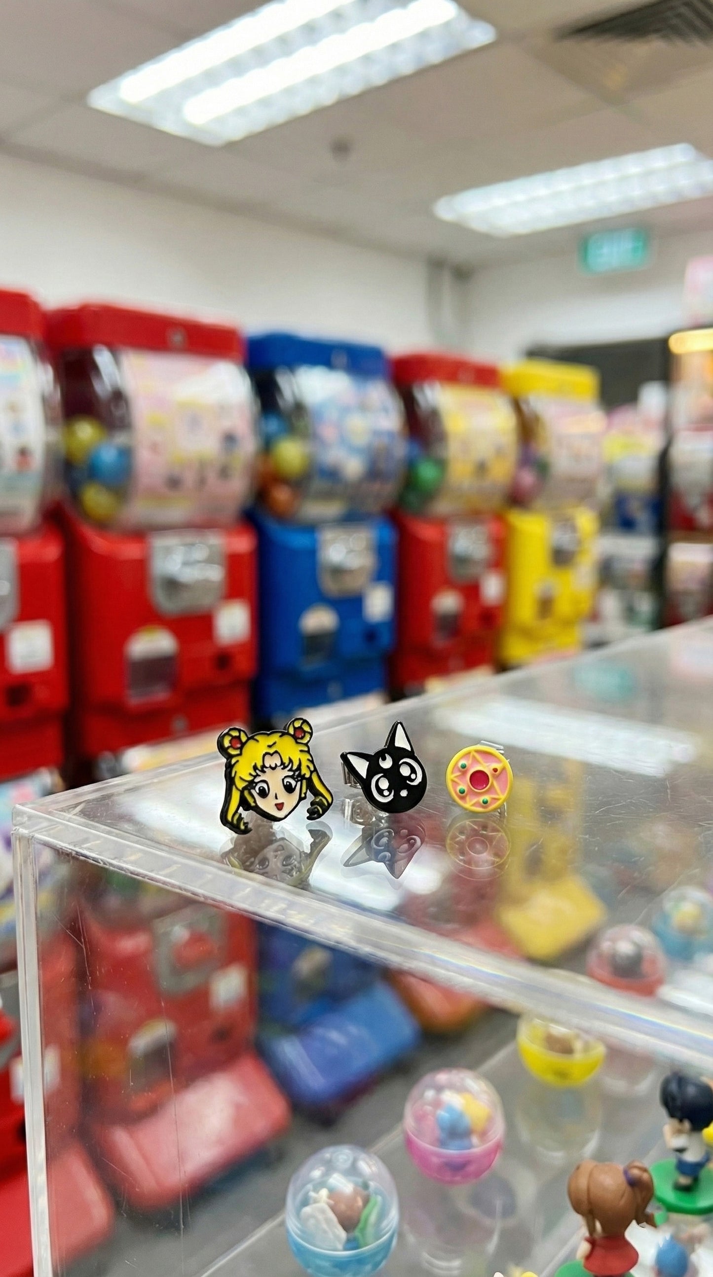 Sailor Moon anime earring set displayed on clear acrylic box with colorful gachapon machines background