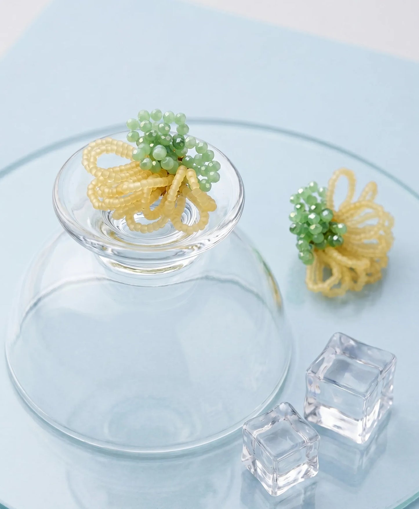 A detailed product shot showing the front and back of mint green and yellow beaded earrings resting on a glass tile with ice cubes. The image highlights the voluminous beadwork and gold-tone stud backing