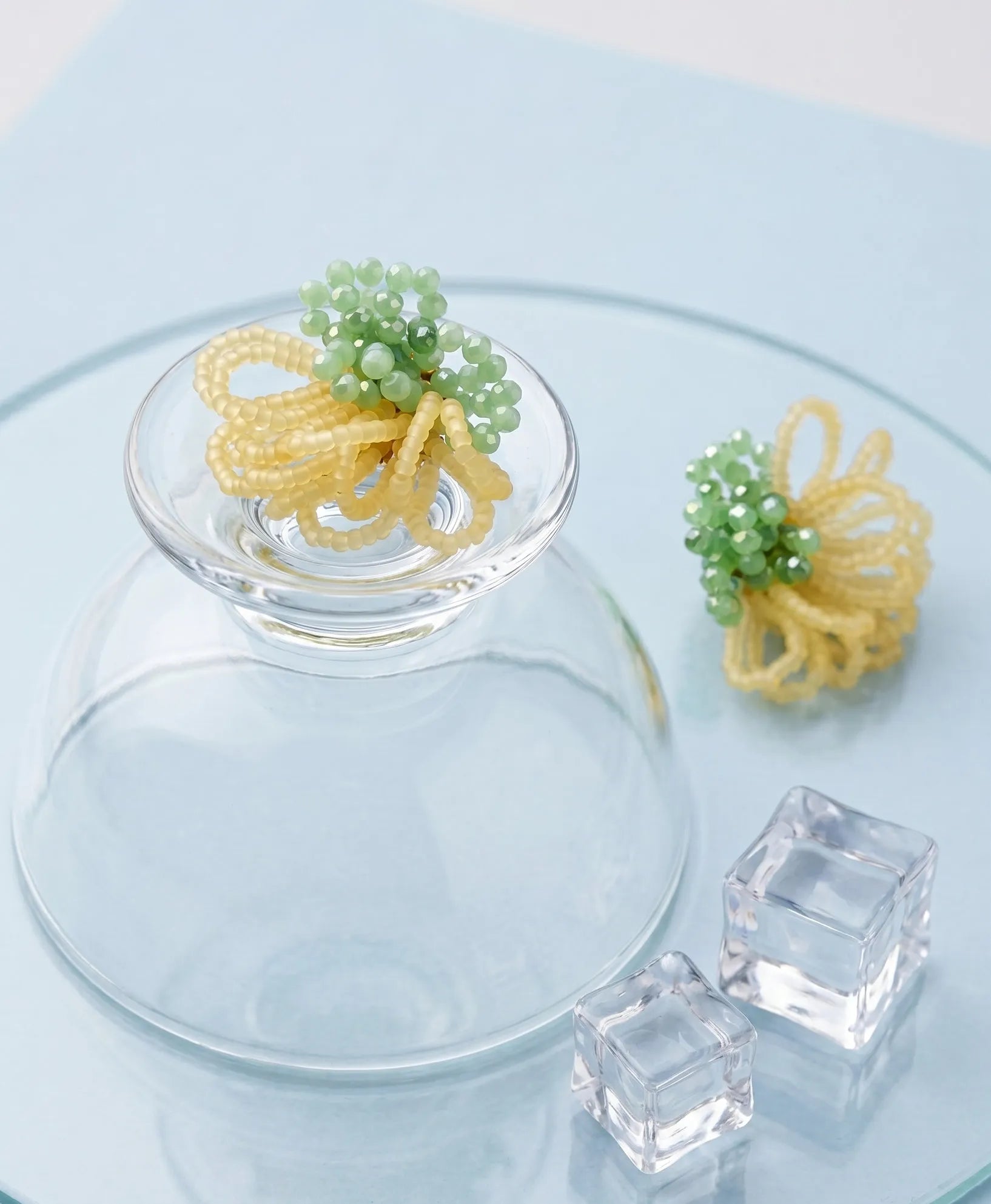 A detailed product shot showing the front and back of mint green and yellow beaded earrings resting on a glass tile with ice cubes. The image highlights the voluminous beadwork and gold-tone stud backing