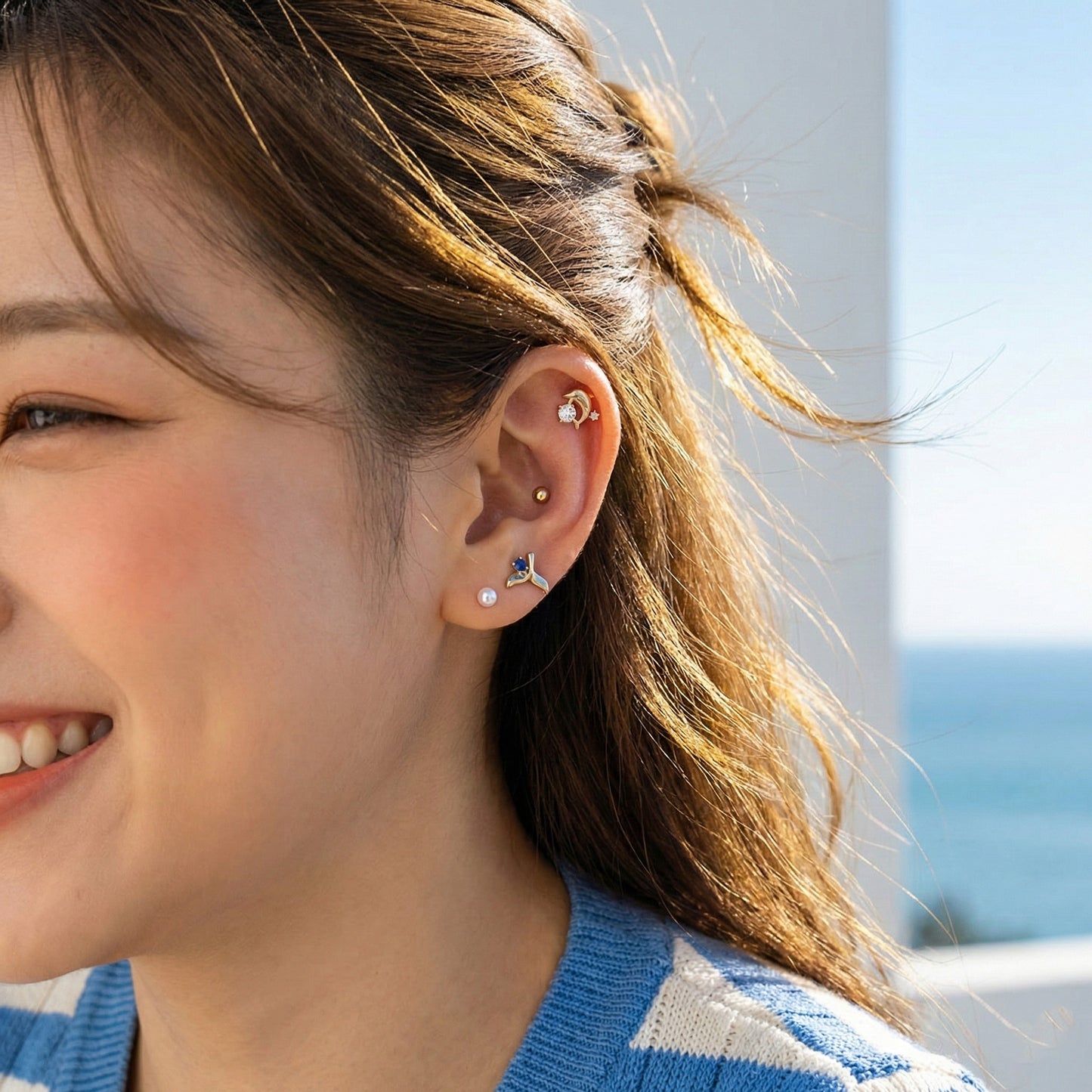 Model wearing layered ear piercings with dolphin and whale tail studs against blue ocean background