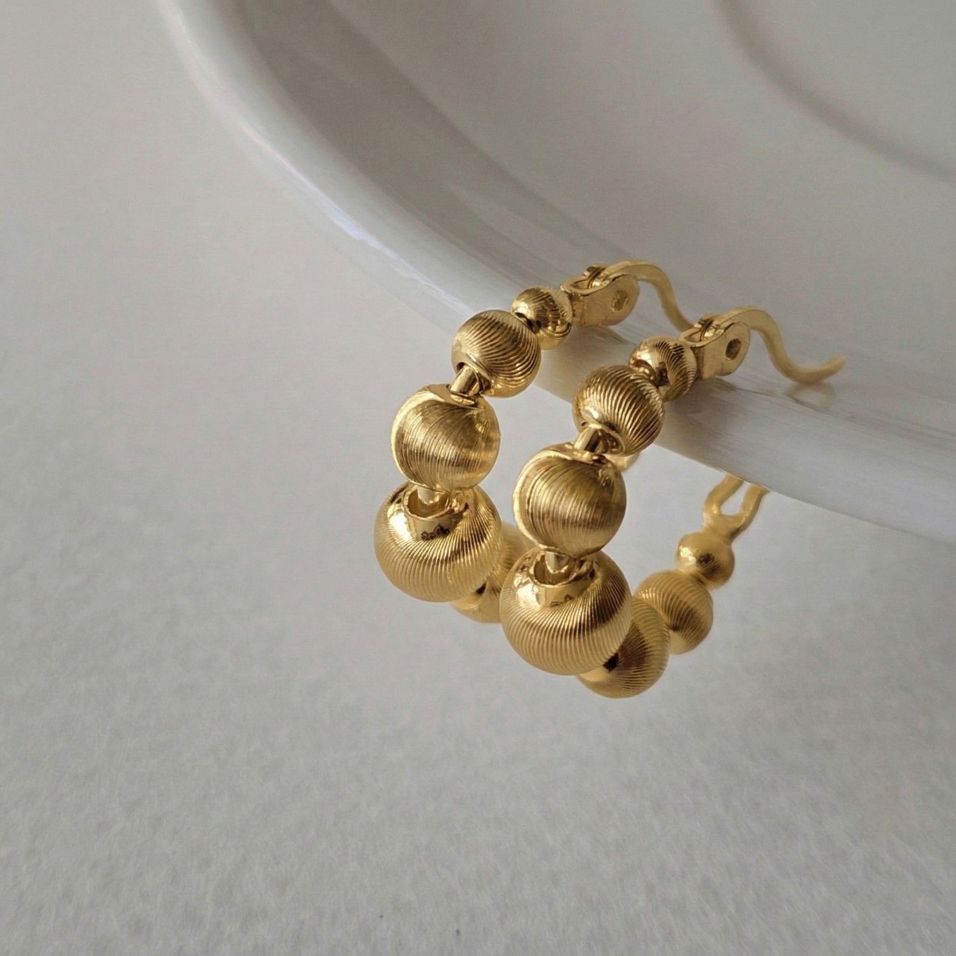 Gold textured sphere hoop earrings on clear display stand, featuring brushed finish graduated bead design with secure hinge closure, gold-plated brass with sterling silver posts, Korean crafted jewelry