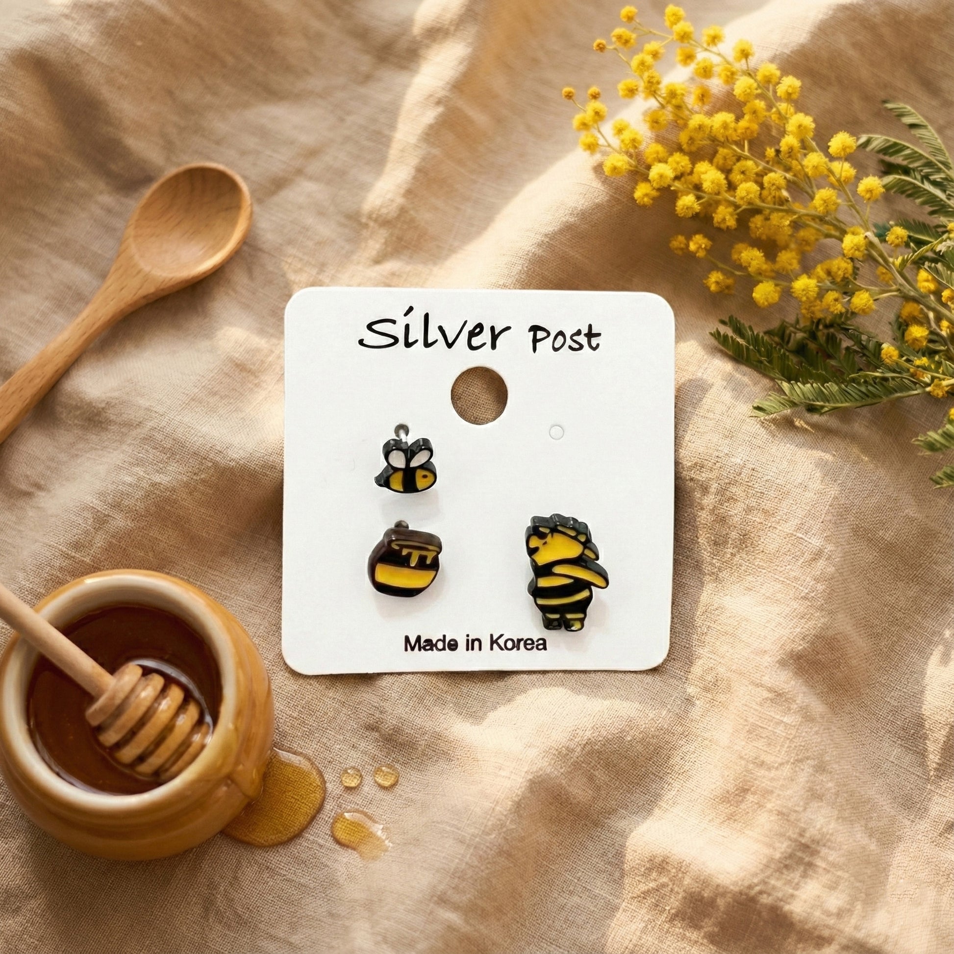 Handmade Pooh, honey pot, and bee earrings on a "Silver Post" card, styled with honey dipper, mimosa flowers, and beige linen.