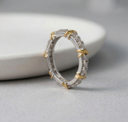 Side-angle product shot of the two-tone 925 silver eternity ring leaning against a white object, clearly showing the full-band cubic zirconia pave and evenly spaced gold X-cross details