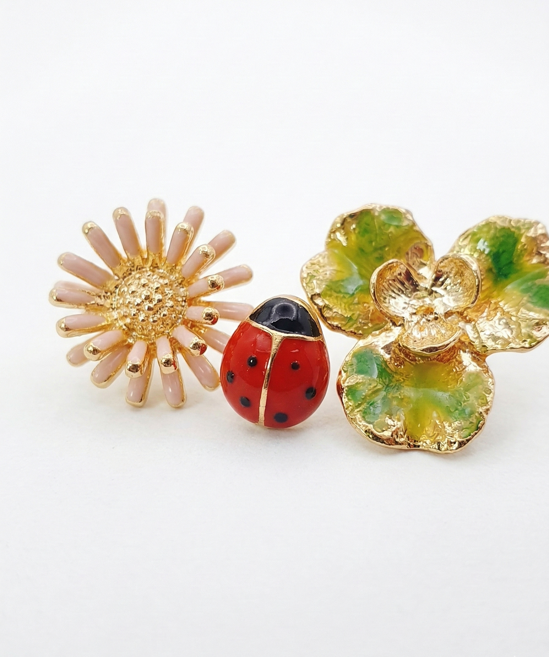 Macro shot of the Retro Red 3-piece earring set: a detailed pink daisy, a red ladybug, and a gold-green clover on a white surface.
