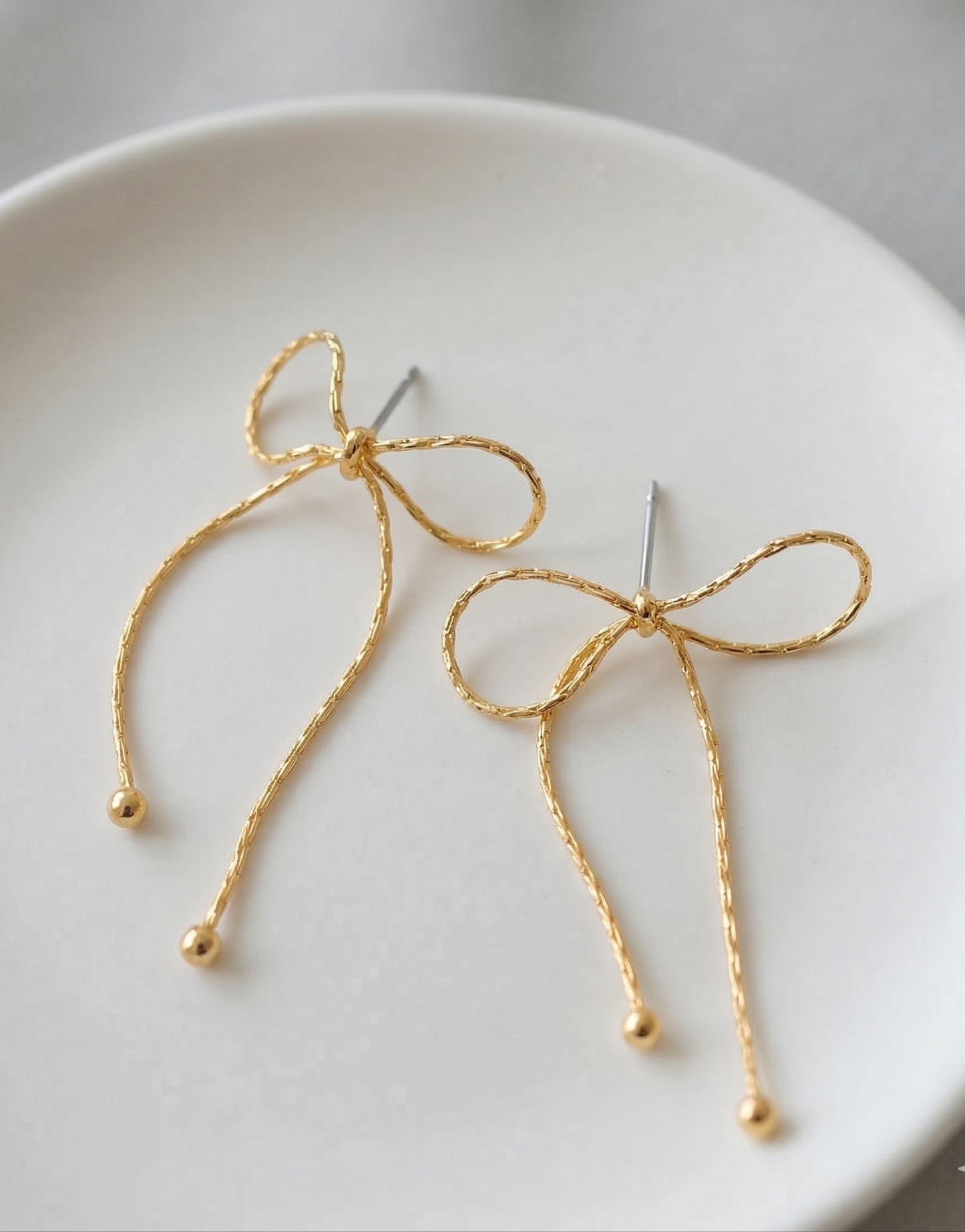 Close-up flat lay of two gold textured chain bow drop earrings on white ceramic plate showing titanium post and ball tip detail