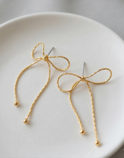 Close-up flat lay of two gold textured chain bow drop earrings on white ceramic plate showing titanium post and ball tip detail