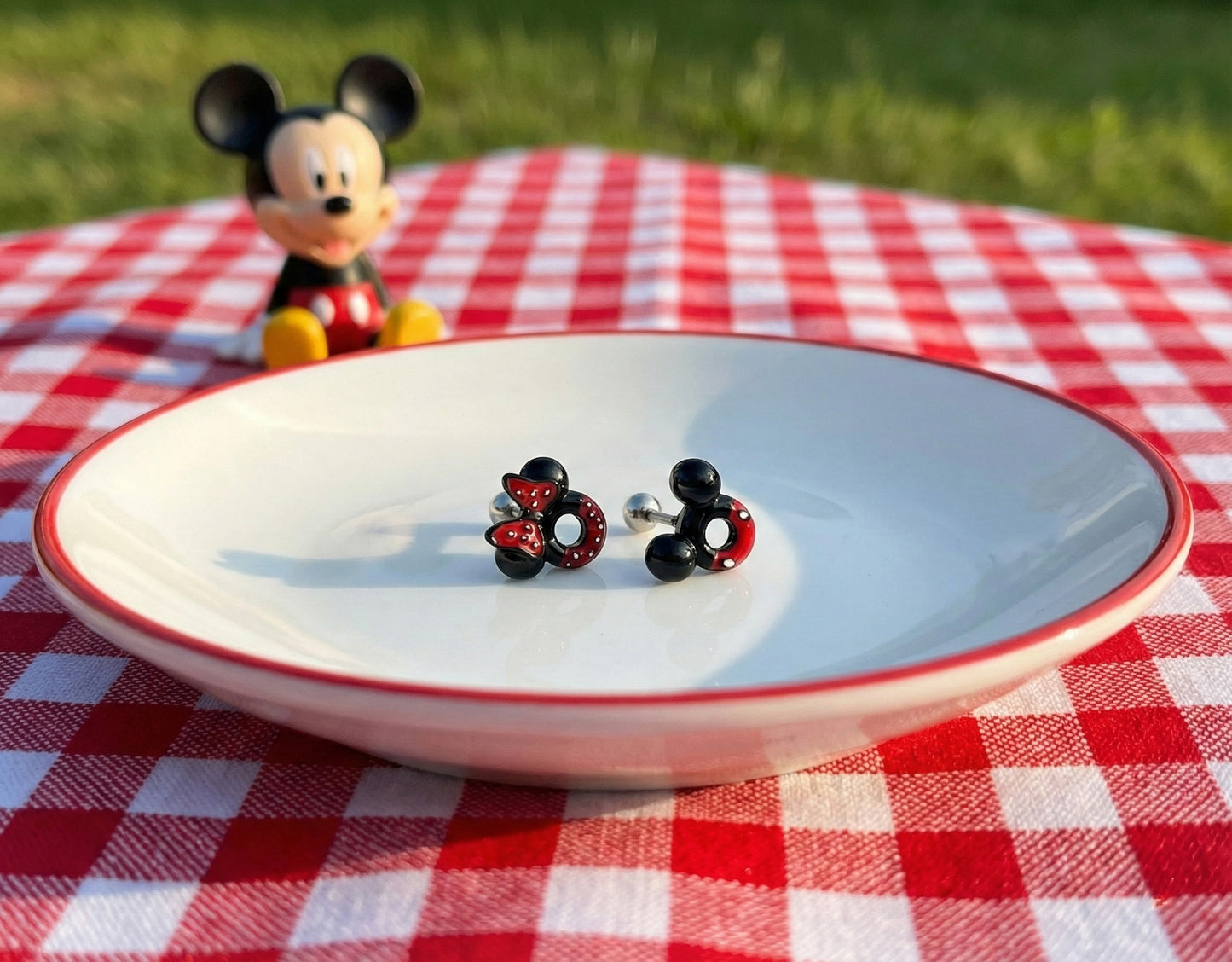 Mickey and Minnie Mouse earrings on a white plate with a Mickey figurine in the background on a red picnic tablecloth