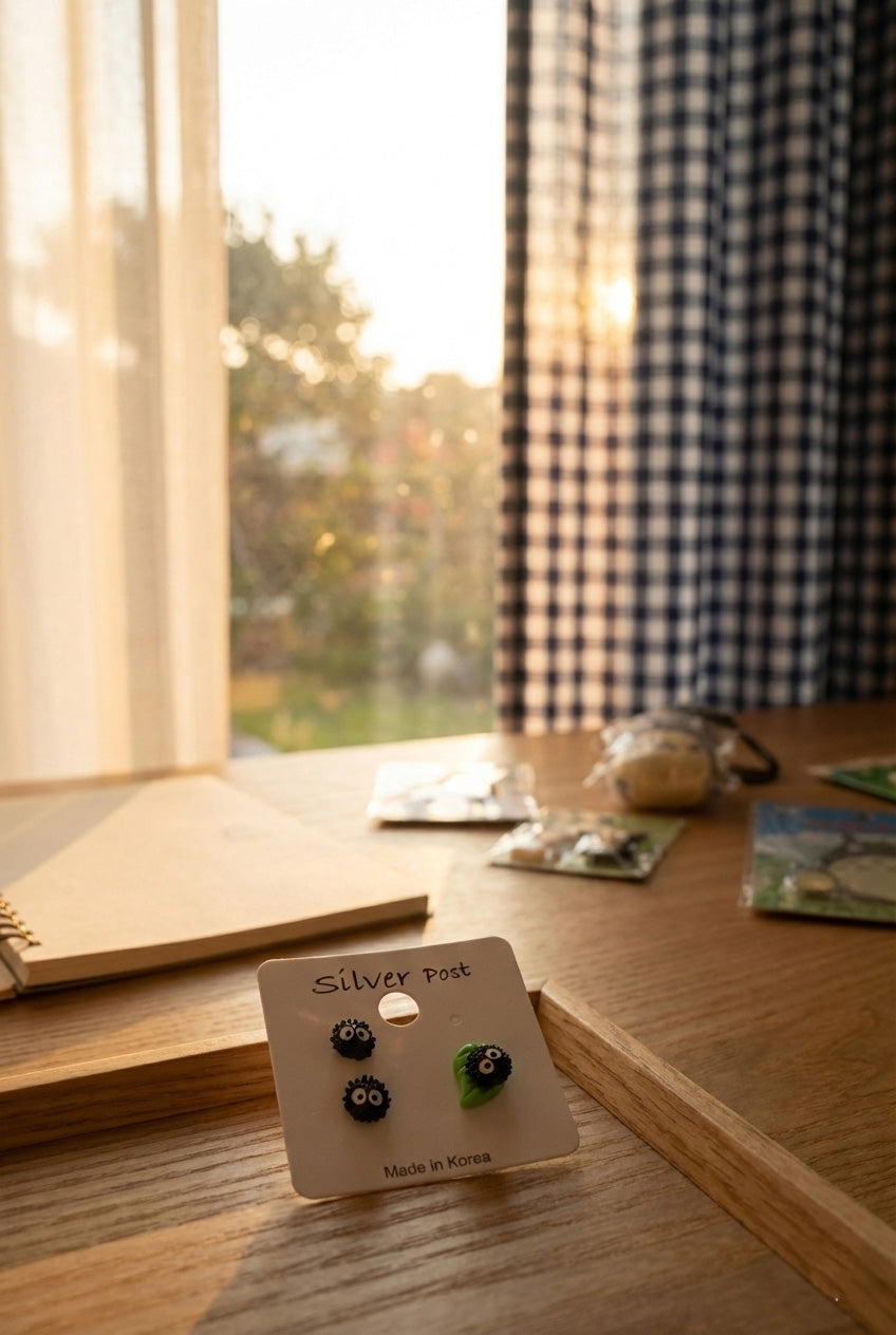 
Soot Sprite earring set on white card with silver post label placed on wooden desk with notebook and Ghibli collectibles
