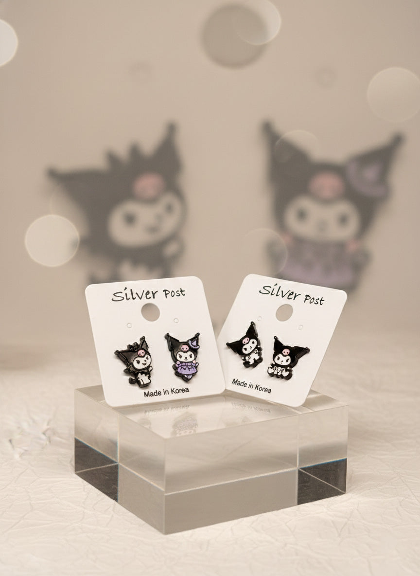 Two pairs of Kuromi-themed enamel earrings on white Silver Post cards, displayed on a transparent acrylic cube with blurred Kuromi figures in the background.