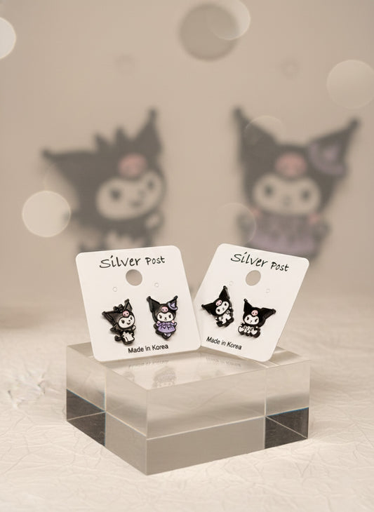 Two pairs of Kuromi-themed enamel earrings on white Silver Post cards, displayed on a transparent acrylic cube with blurred Kuromi figures in the background.