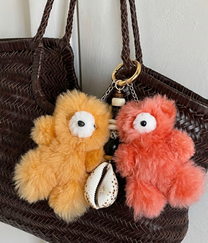 Orange and coral alpaca bear keyrings hanging on a dark woven straw tote bag with cowrie shell and beaded charm accent
