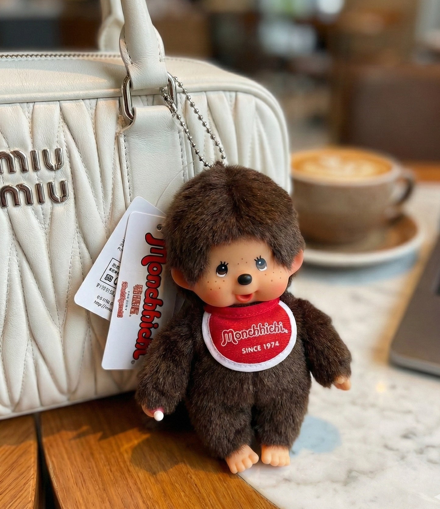  Monchhichi Classic Boy plush bag charm clipped to white Miu Miu matelassé leather handbag at café with latte in background