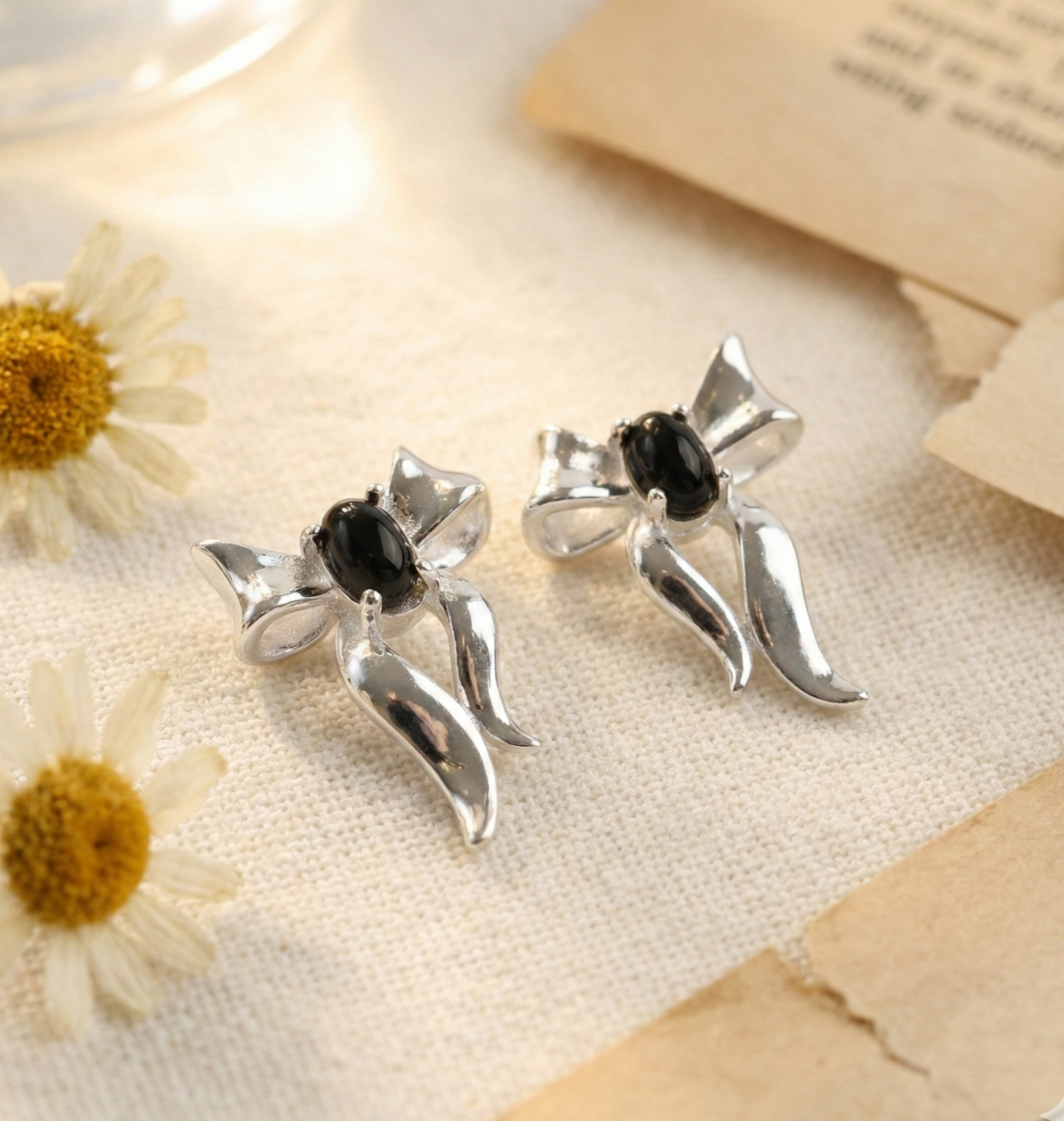 Pair of silver bow earrings with black agate center laid on linen fabric with white daisy flowers — vintage aesthetic flat lay