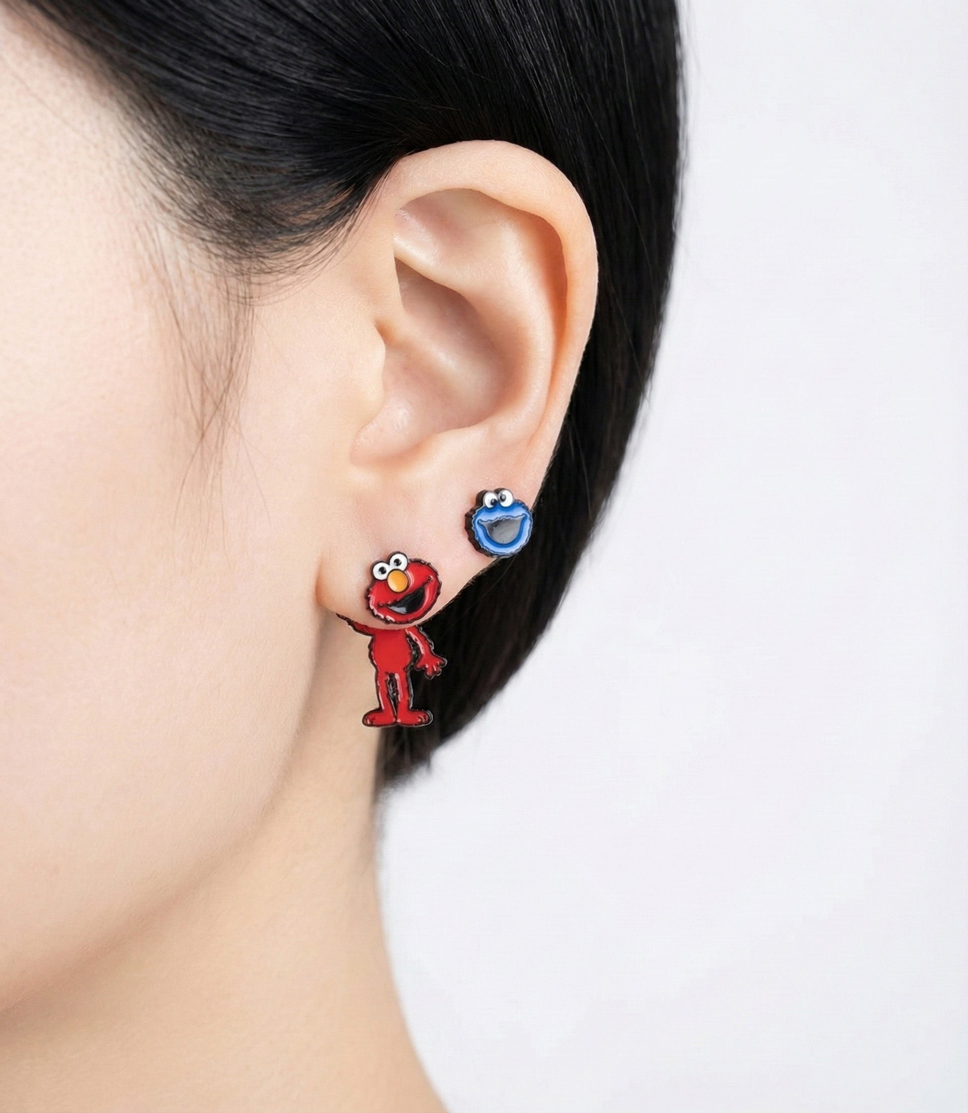 Elmo and Cookie Monster mismatched earrings styled on ear; Elmo full body dangles below, Cookie Monster head sits on lobe.