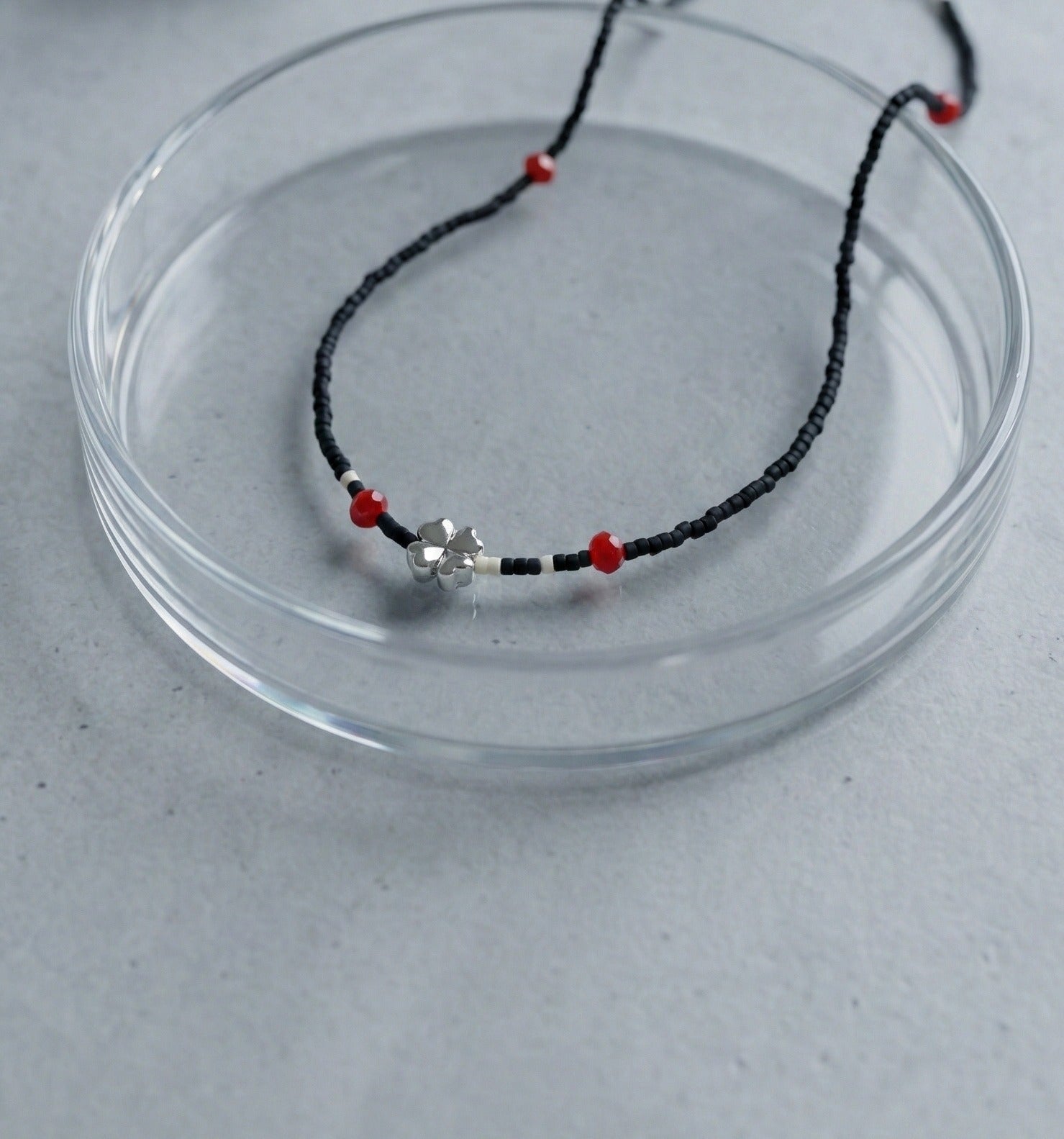 Detail shot of black seed bead choker necklace with silver clover pendant and red accent beads on glass plate