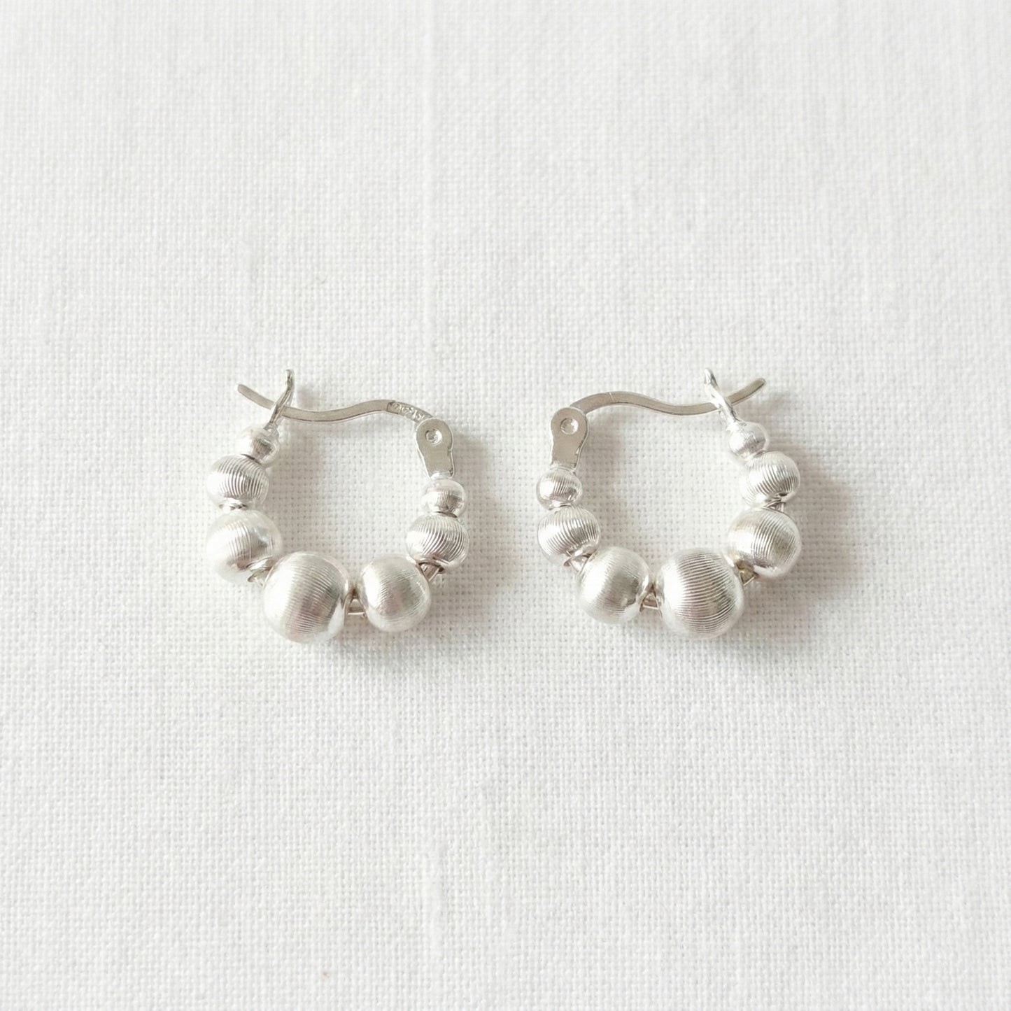 Pair of silver beaded hoop earrings on white linen background, displaying textured spherical beads in graduated sizes, 925 sterling silver posts hypoallergenic, lightweight comfortable fit for sensitive ears