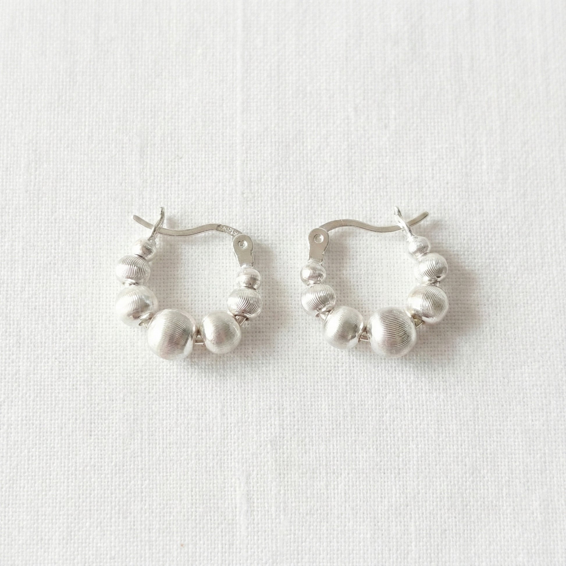 Pair of silver beaded hoop earrings on white linen background, displaying textured spherical beads in graduated sizes, 925 sterling silver posts hypoallergenic, lightweight comfortable fit for sensitive ears