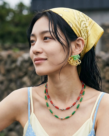 A woman radiating effortless K-summer fashion, wearing a yellow bandana, a pastel polka-dot slip dress, layered beaded necklaces, and mint green beaded earrings, set against a rustic stone wall.