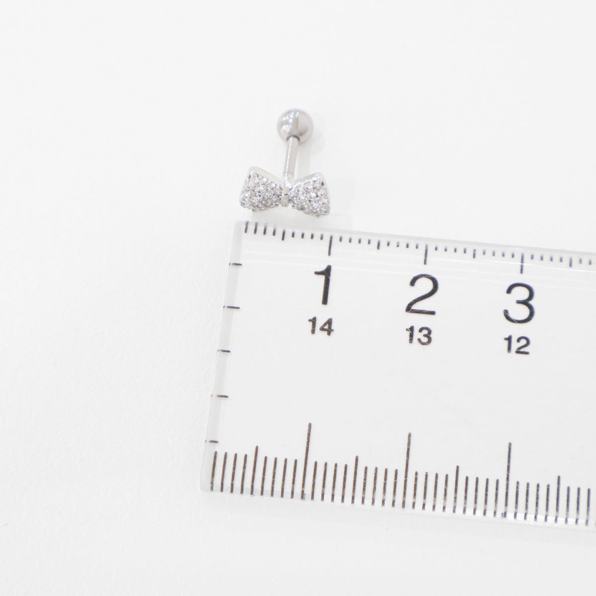 Silver bow barbell earring placed next to a ruler, showing approximate width of 1.3 cm