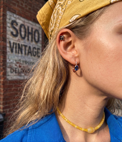 Side profile of a model showcasing a deep blue oval sapphire-like earring in a bezel setting and a black crystal clover outer conch piercing. She wears a yellow beaded choker necklace and a yellow bandana, with a rustic brick wall and a "SOHO VINTAGE" sign blurred in the background.