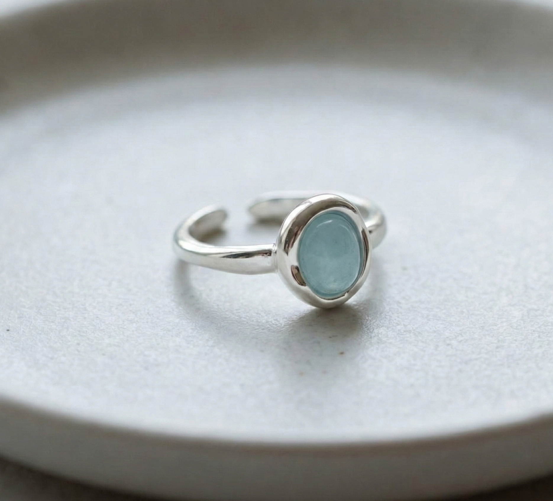 Close-up of 925 sterling silver natural aquamarine adjustable open ring with oval cabochon bezel setting and smooth band on white ceramic surface showing aqua blue stone clarity