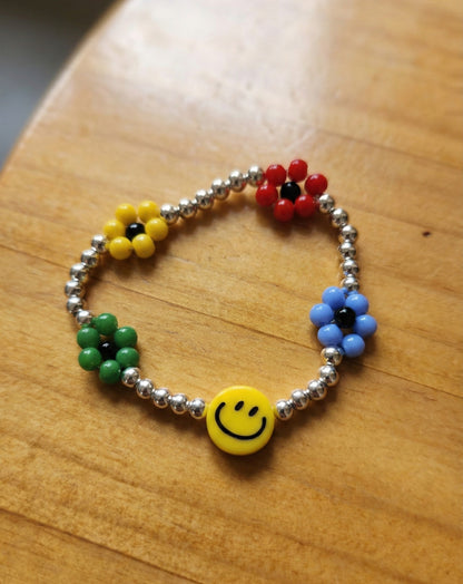 Smiley face yellow bead bracelet with four colorful seed bead flowers in red, yellow, green, and blue on wooden surface