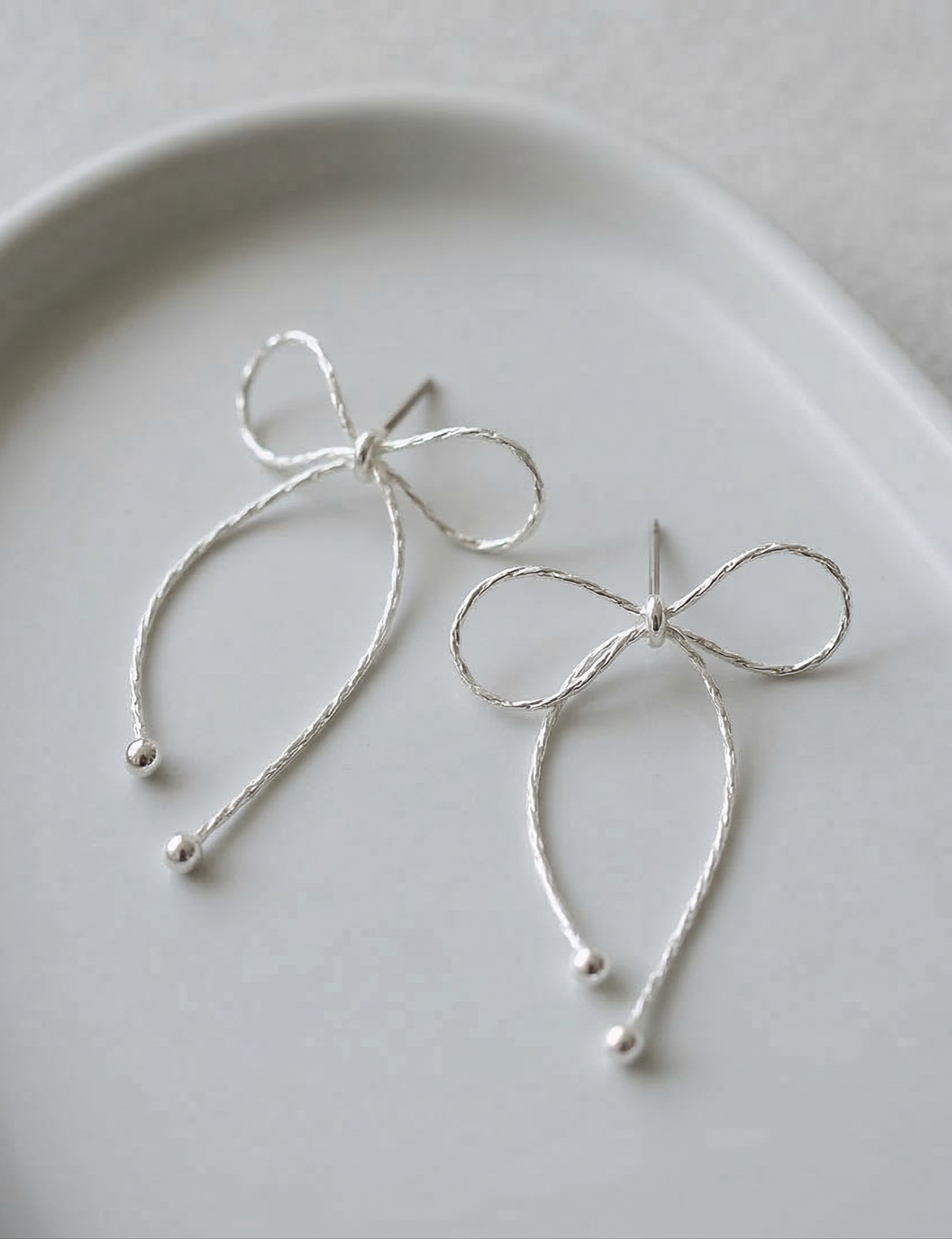 Close-up flat lay of two silver textured chain bow drop earrings on white ceramic plate showing delicate chain texture and ball tip finish