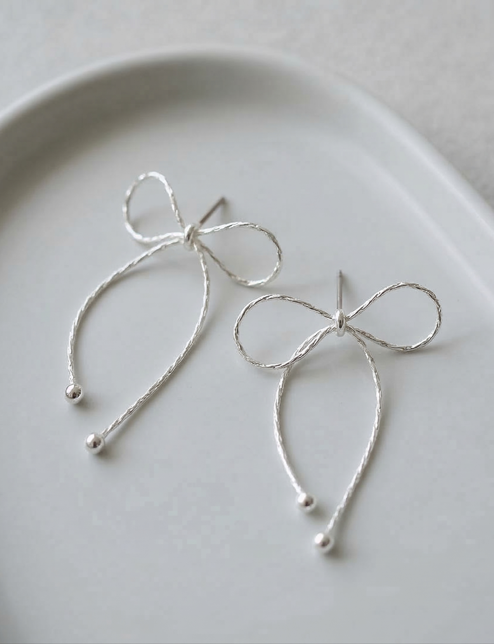 Close-up flat lay of two silver textured chain bow drop earrings on white ceramic plate showing delicate chain texture and ball tip finish