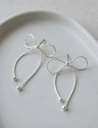 Close-up flat lay of two silver textured chain bow drop earrings on white ceramic plate showing delicate chain texture and ball tip finish