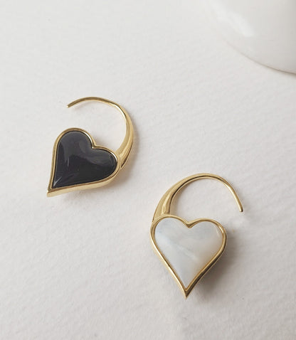 Two heart hook earrings in gold, one with black onyx and one with white mother-of-pearl, laid flat on textured white paper.