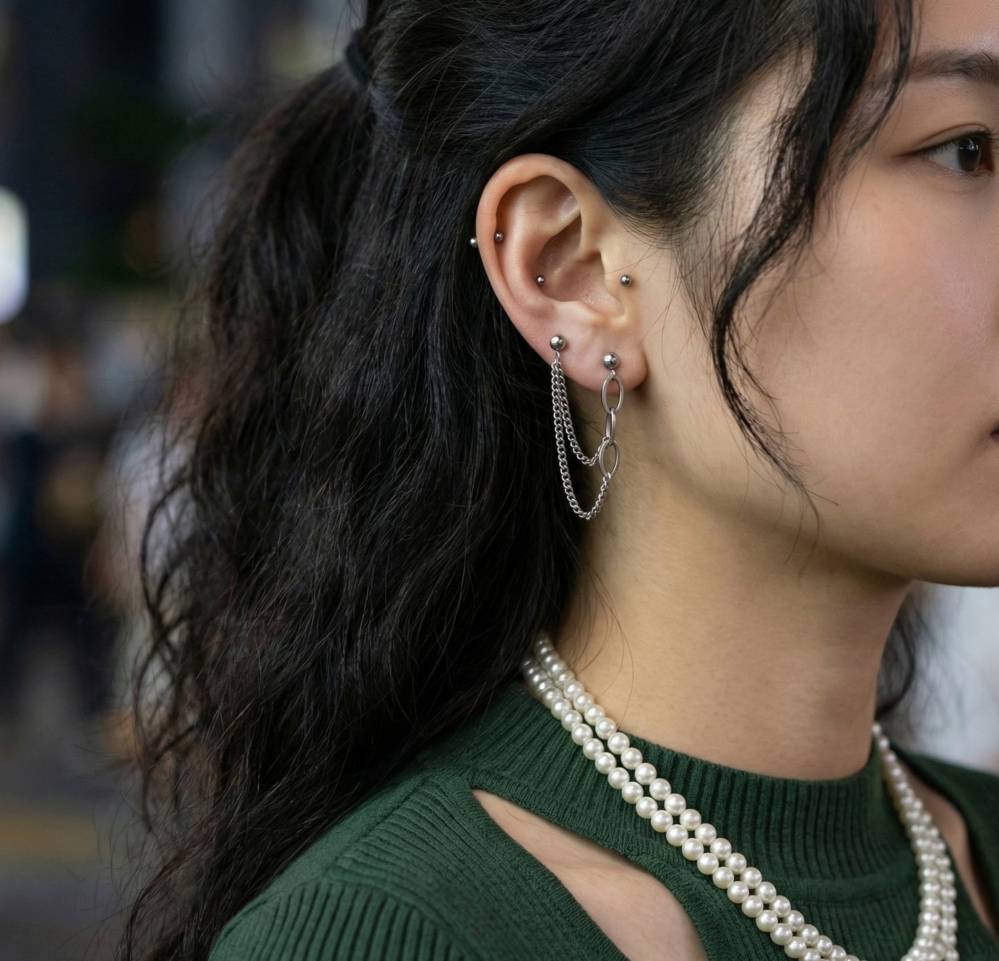 "Woman wearing double piercing chain earrings with green knit sweater and pearl necklace for layered Y2K style"