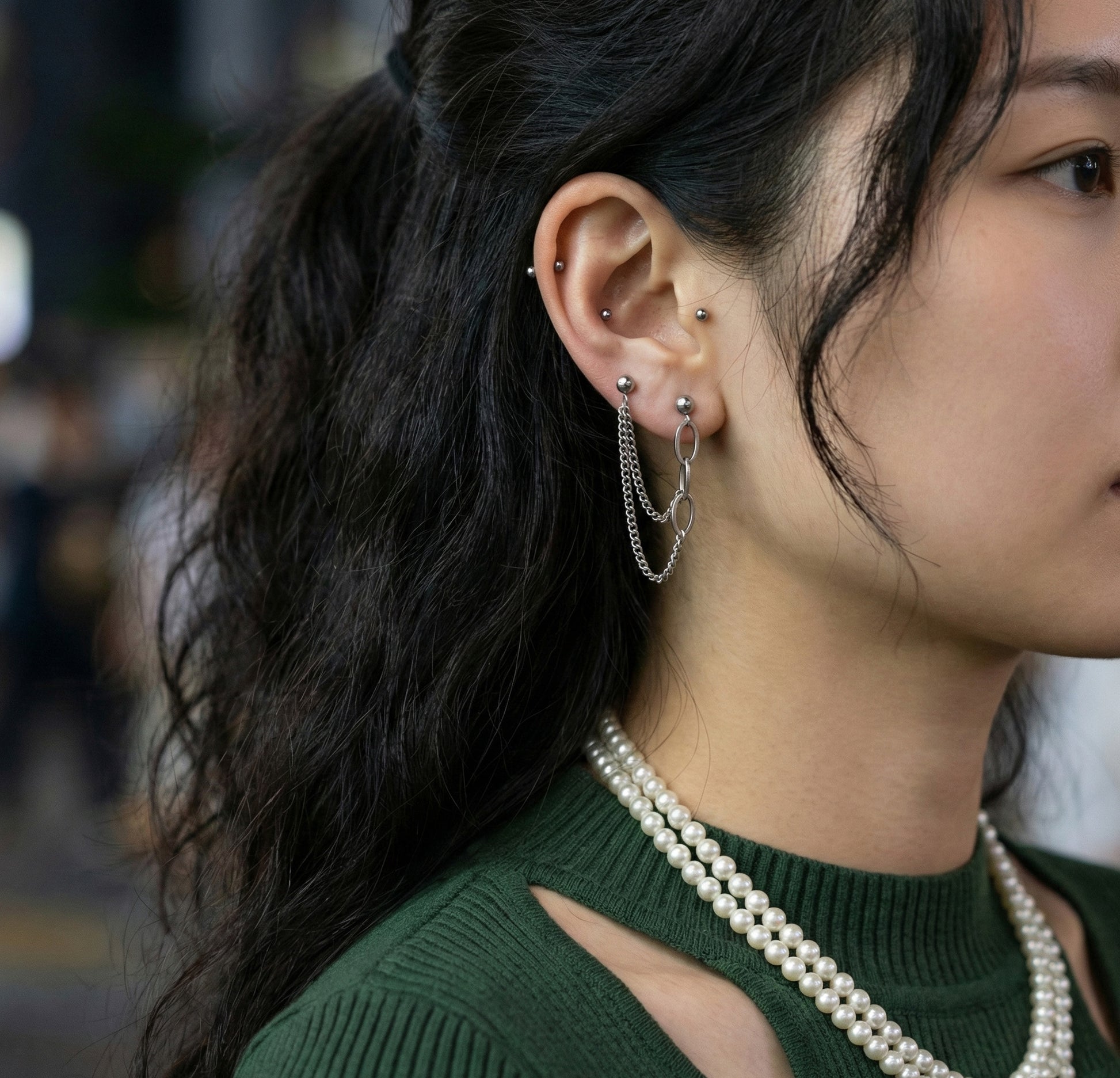 "Woman wearing double piercing chain earrings with green knit sweater and pearl necklace for layered Y2K style"