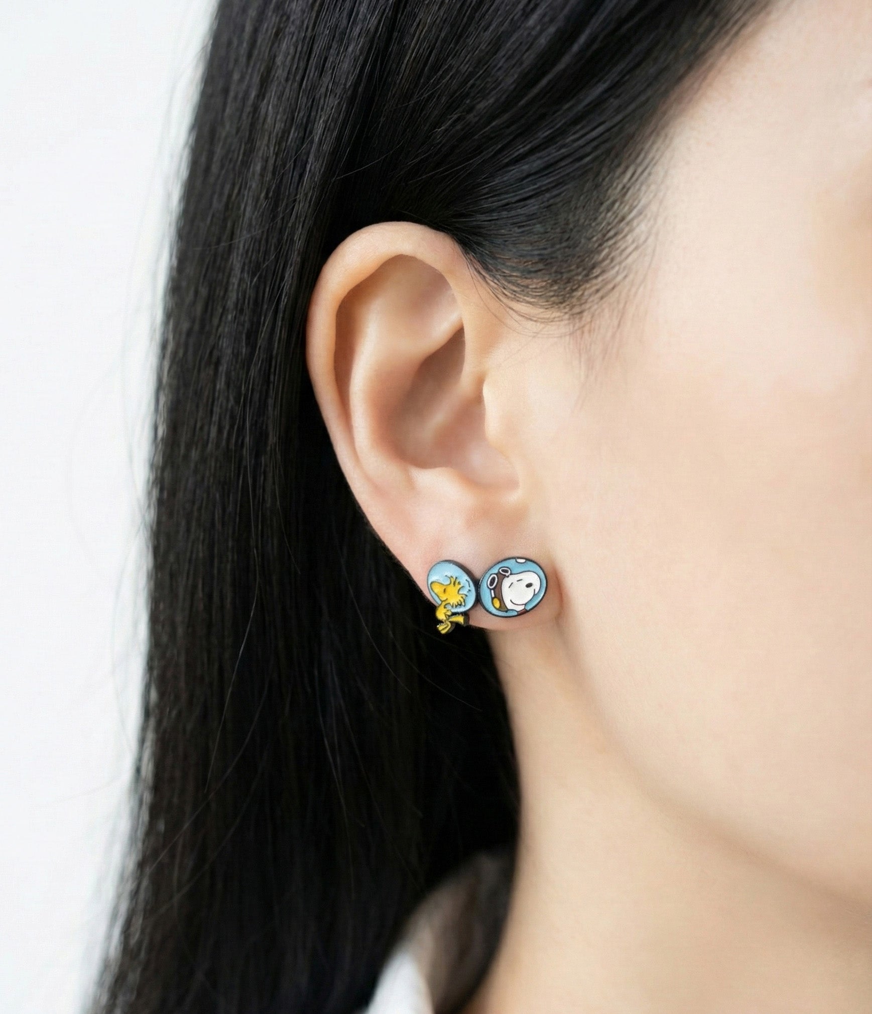 Model wearing mismatched Snoopy and Woodstock earrings — both characters in astronaut helmets, styled together on one ear.