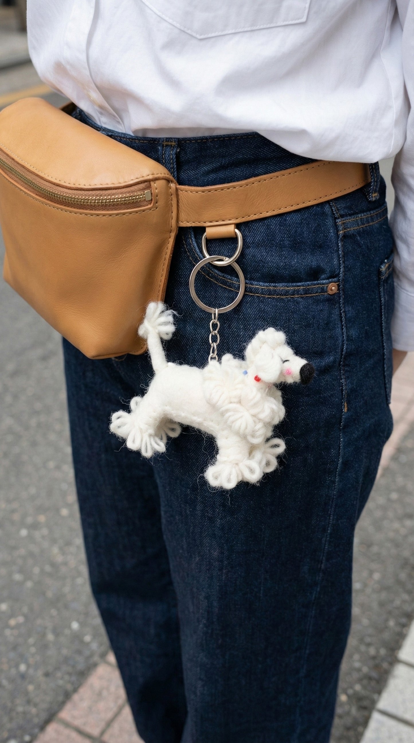 Wool felt poodle keychain hanging from a tan waist bag worn with white shirt and denim jeans