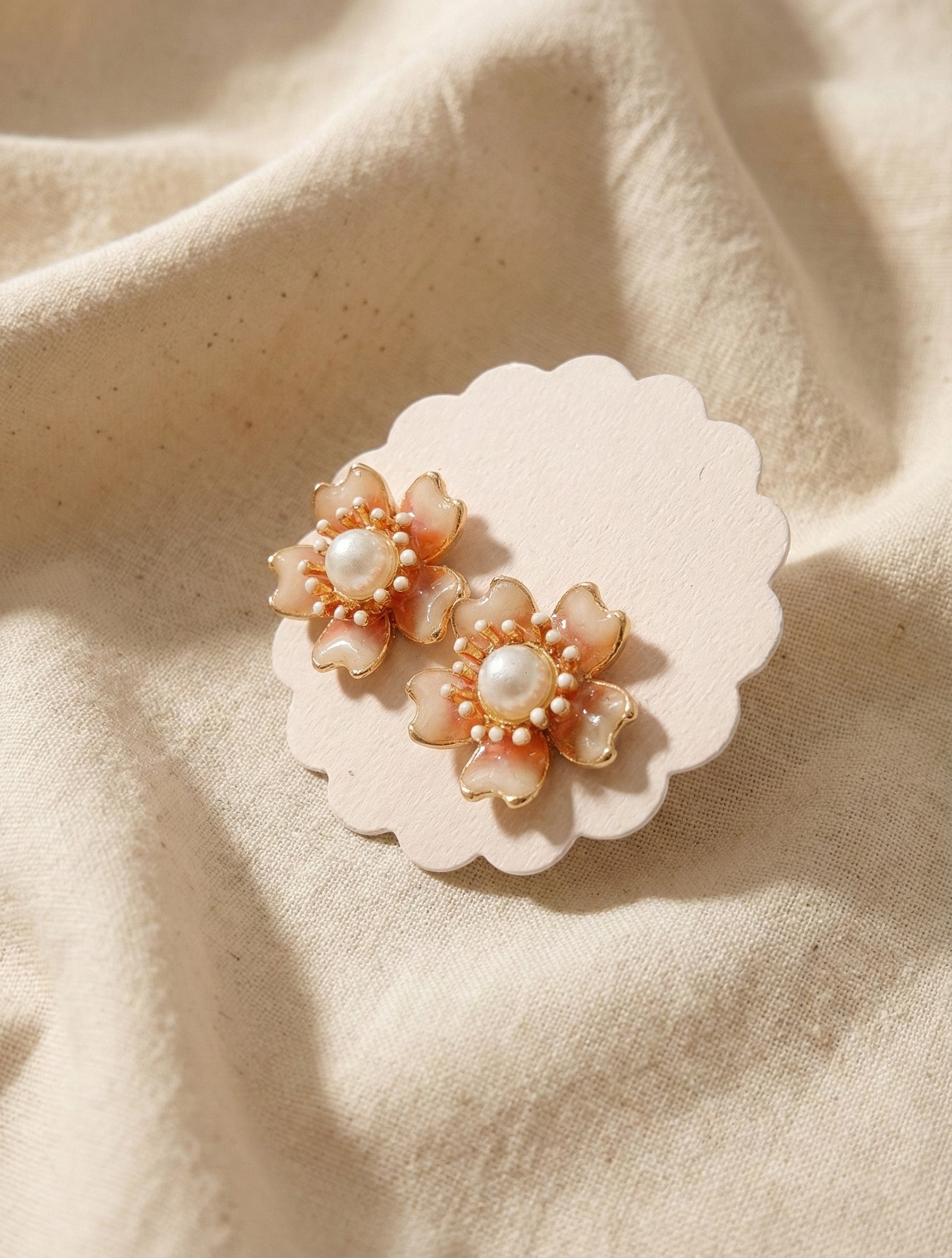 Handcrafted floral stud earrings in warm peach tone with metallic gold rim and pearl, styled on neutral fabric