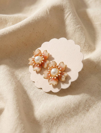 Handcrafted floral stud earrings in warm peach tone with metallic gold rim and pearl, styled on neutral fabric