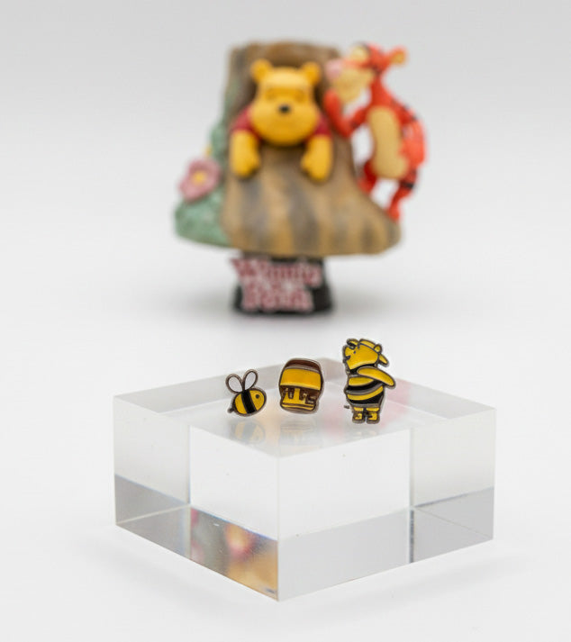 Bee, honey pot, and Winnie the Pooh earrings standing on a clear acrylic block with a blurred Pooh and Tigger figure in the background.