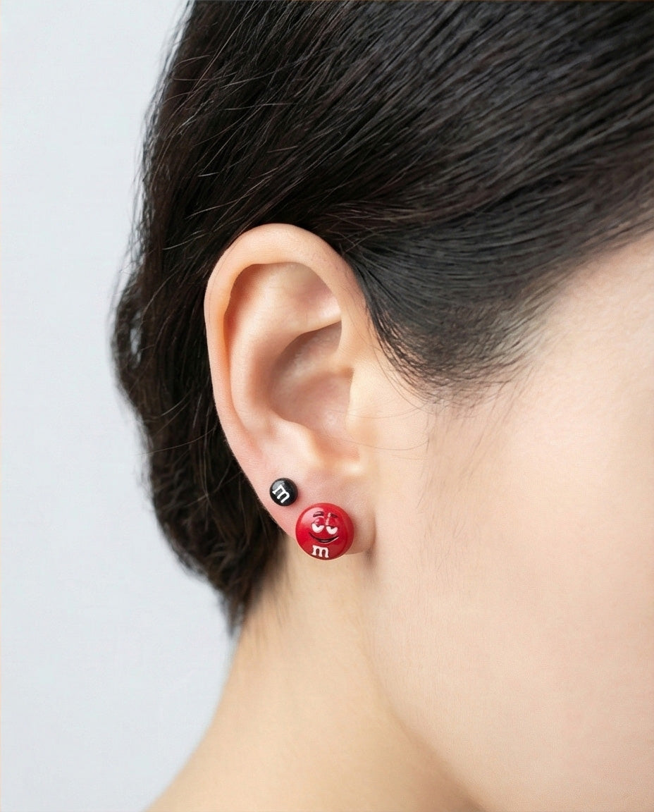 Woman wearing M&M's candy earrings featuring red character with smile and black mini stud, side profile showcasing fun double piercing combination, lightweight enamel jewelry, 925 silver posts made in Korea