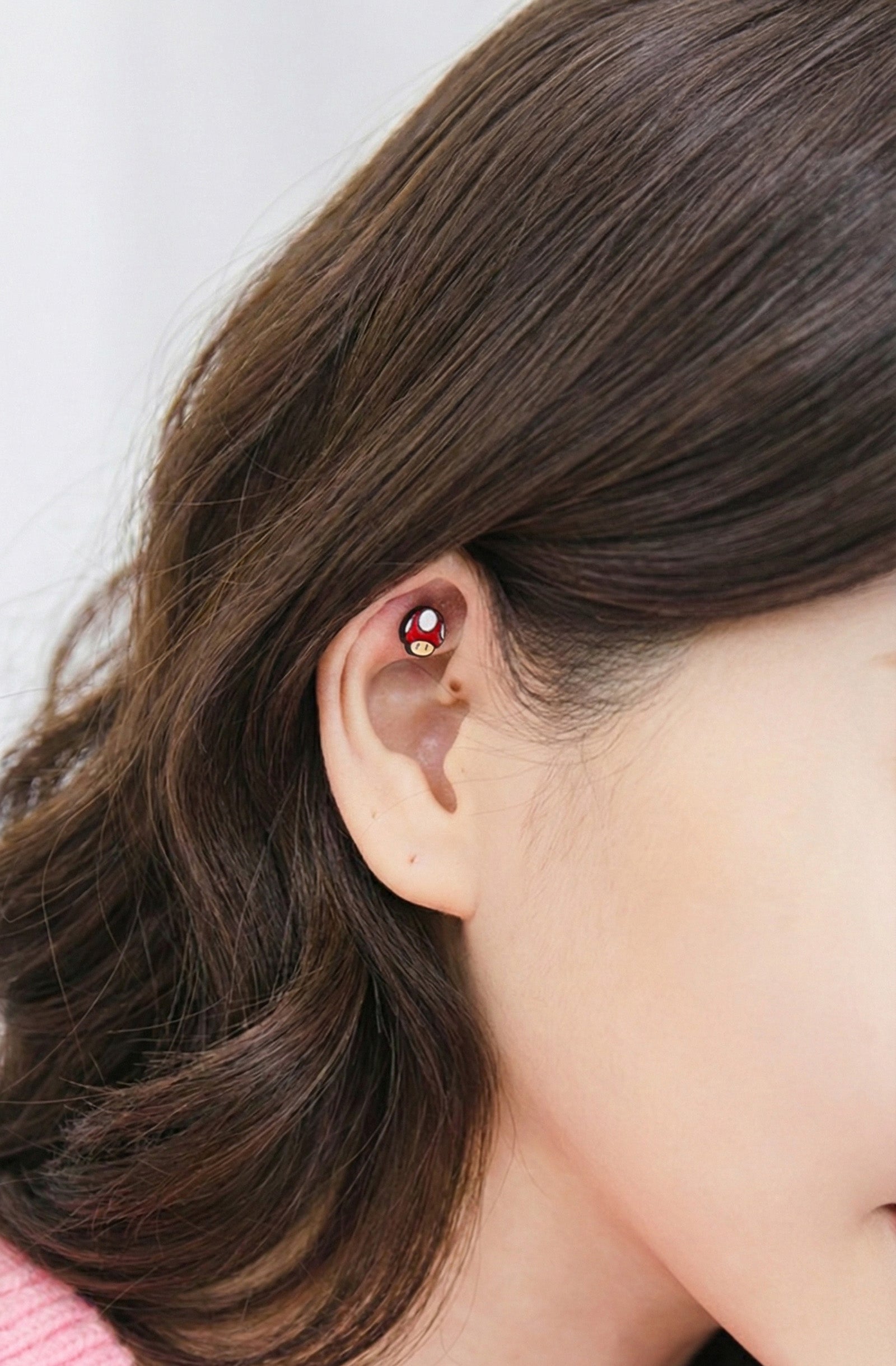 Red mushroom stud piercing worn on the inner ear fold of a model with brown hair.