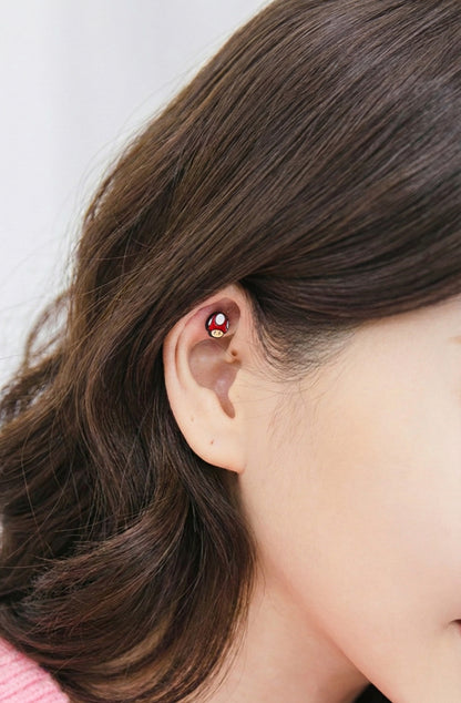 Red mushroom stud piercing worn on the inner ear fold of a model with brown hair.