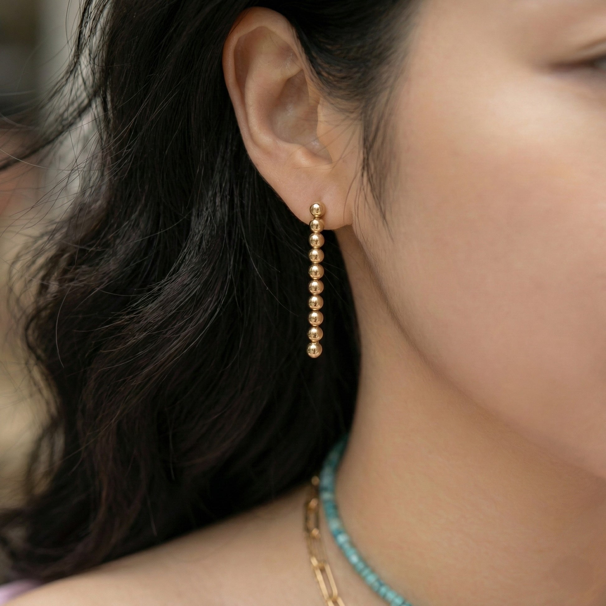 Side view of woman wearing gold ball drop earrings with turquoise necklace